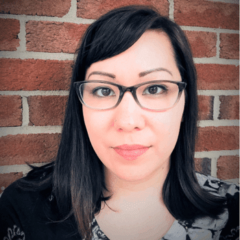 a woman wearing glasses standing in front of a brick wall.